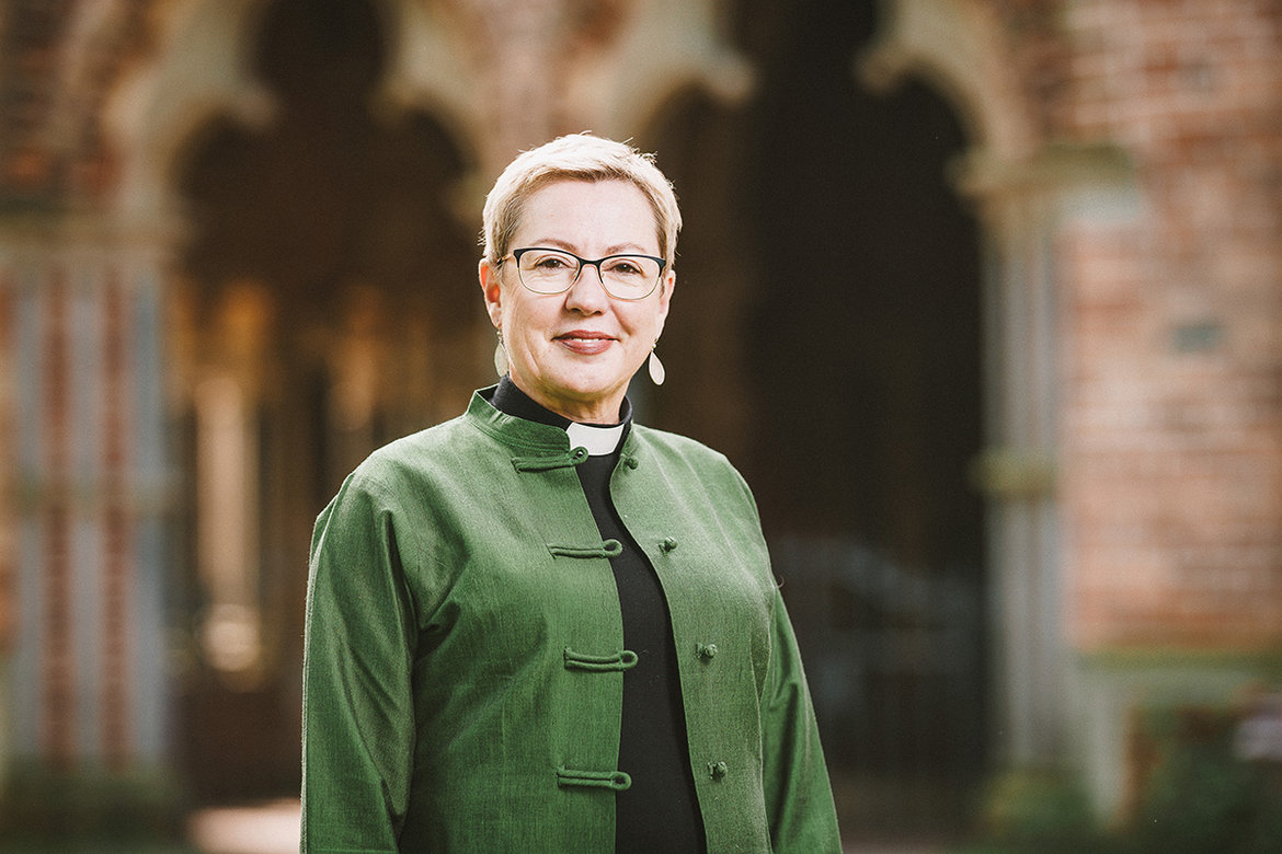 Pröpstin Petra Kallies steht vor einem Torbogen des Doms zu Lübeck. Sie trägt eine grüne Jacke, eine Brille und hat kurzes blondes Haar und lächelt in die Kamera